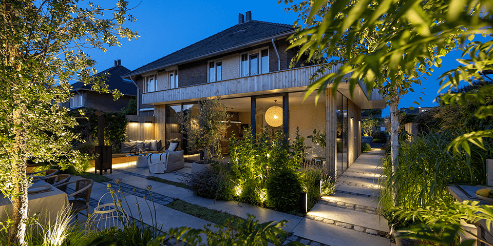 In einem beleuchteten Garten mit Terrasse sind unterschiedliche Formen direkter und indirekter Gartenbeleuchtung zu sehen: Außenwandleuchten, Outdoor-Hängeleuchten, LED-Streifen, Wegeleuchten, Bodeneinbauleuchten und Gartenstrahler. 