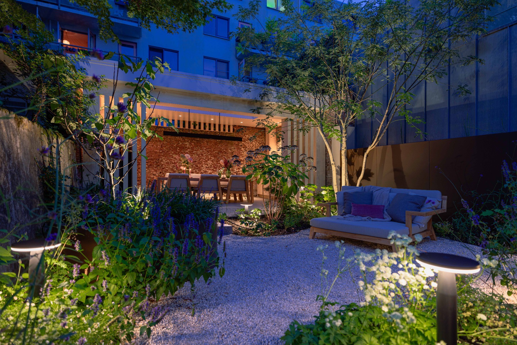 Ein moderner Garten mit Bepflanzung, einer gemütlichen Sitzecke und einem Pool wird von verschiedenen Außenleuchten erhellt. Im Mittelpunkt stehen die Zaun-, Baum- und Beetbeleuchtung.