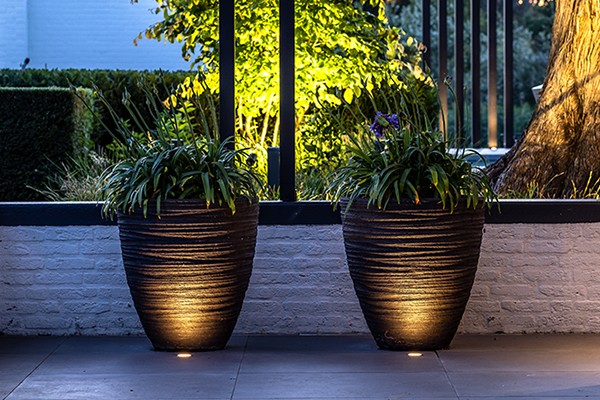 Bodenleuchten auf einer Terrasse werfen ein dekoratives Licht auf zwei Pflanzkübel. Im Hintergrund ist ein beleuchteter Garten zu sehen.