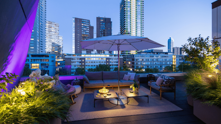 Eine moderne Dachterrasse in einer Großstadt wird mithilfe von exklusiver smarter in-lite-Außenbeleuchtung und intelligentem System-Zubehör in farbiges Licht getaucht. Zu sehen sind eine beleuchtete Lounge-Ecke mit Sonnenschirm, mehrere beleuchtete Grünpf