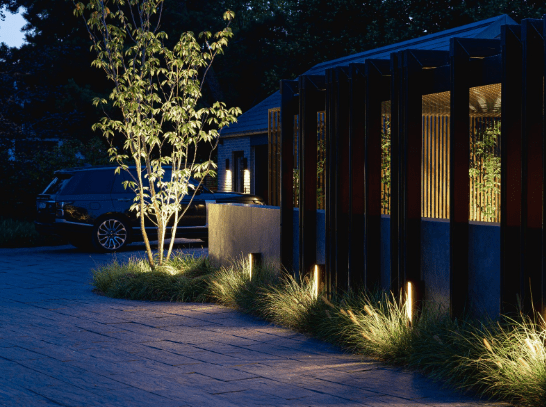 Neben der Zufahrt eines Hauses ist ein Weg zu sehen, der in der Abenddämmerung von mehreren stilvollen Pollerleuchten ausgeleuchtet wird. 