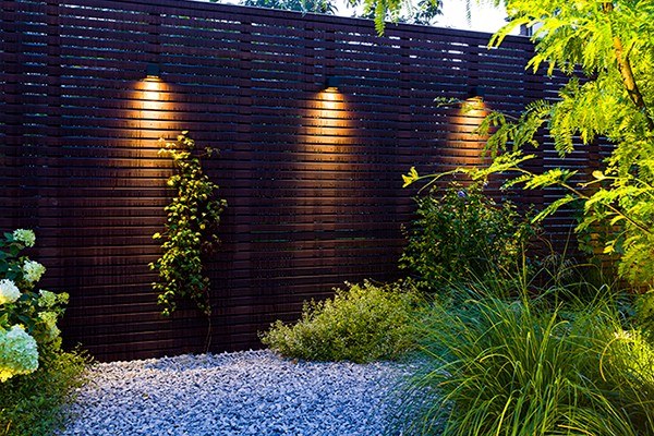 Ein Holz-Sichtschutzzaun, der von drei Außenwandleuchten stimmungsvoll beleuchtet wird.